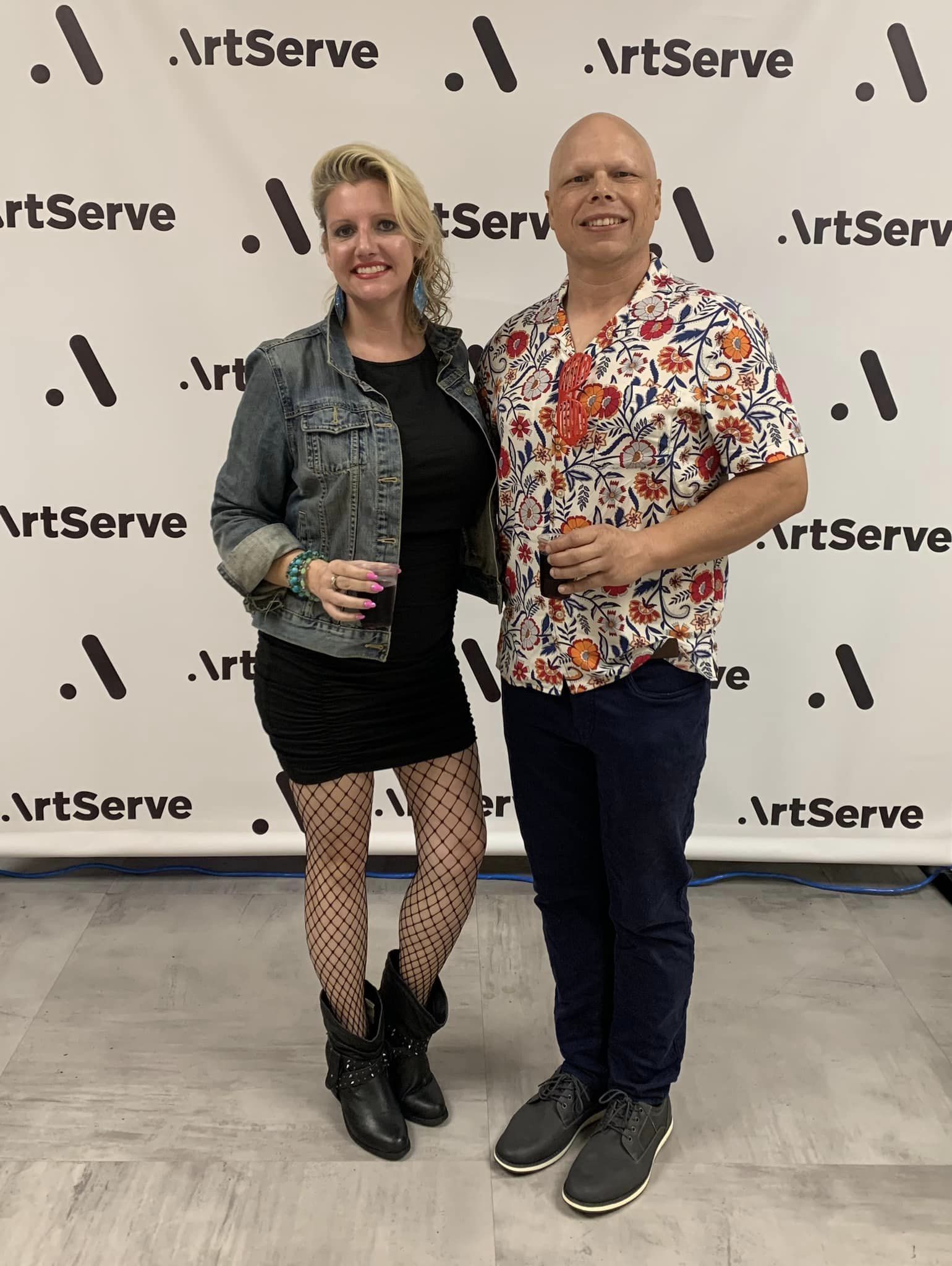 A woman and a man stand smiling in front of an ArtServe backdrop, embodying the essence of arts and culture. The woman wears a denim jacket, black dress, and fishnet stockings, while the man sports a floral shirt with blue pants. Both are holding drinks, celebrating creativity.