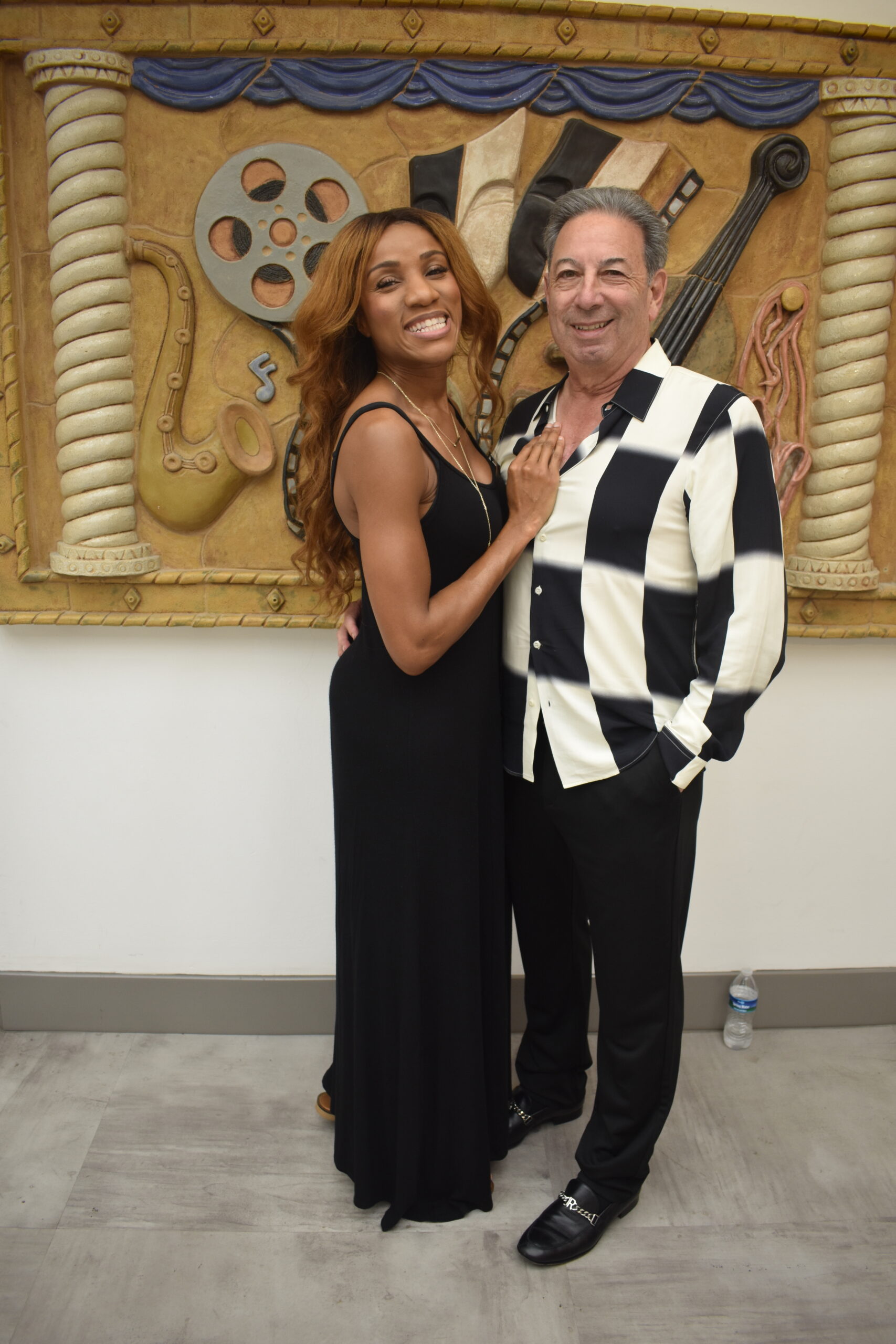 A woman in a black dress and a man in a black and white checkered shirt are posing together, embodying the spirit of arts and culture. They smile warmly in front of a decorative wall adorned with musical and film-themed artwork. A water bottle rests on the floor nearby.