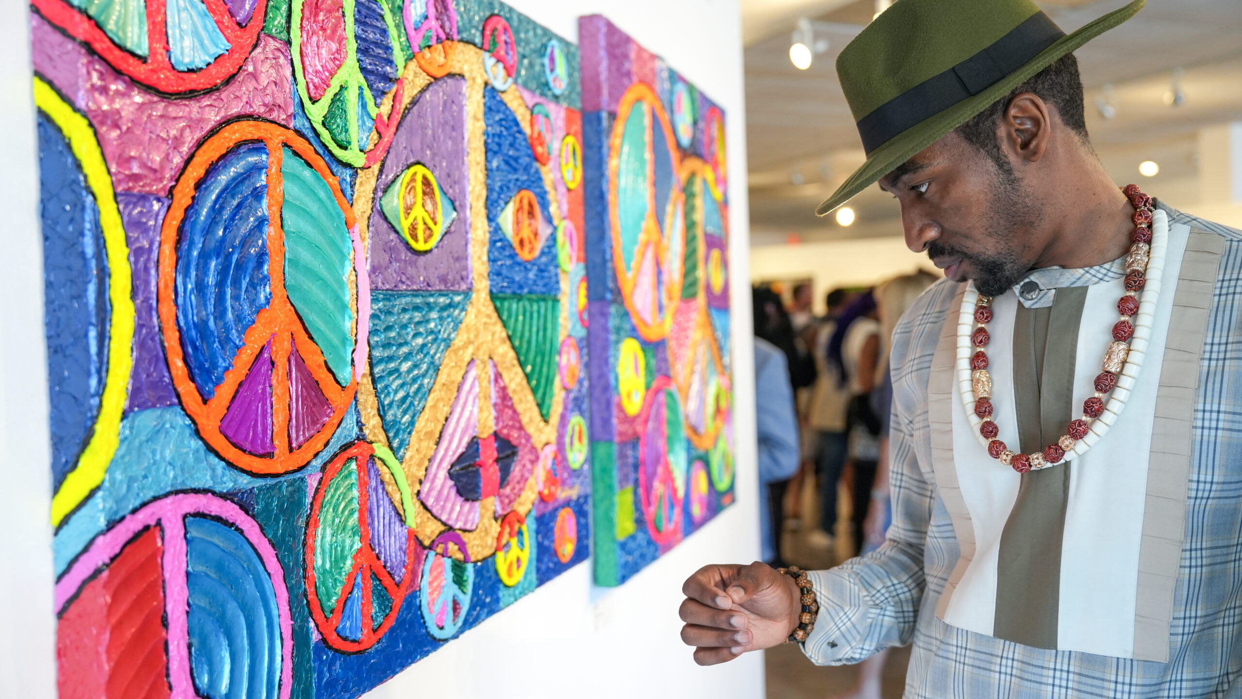 A person wearing a green hat and beaded necklace examines colorful peace symbol artwork in a gallery. The painting features vibrant circles with peace signs. Other people are blurry in the background.